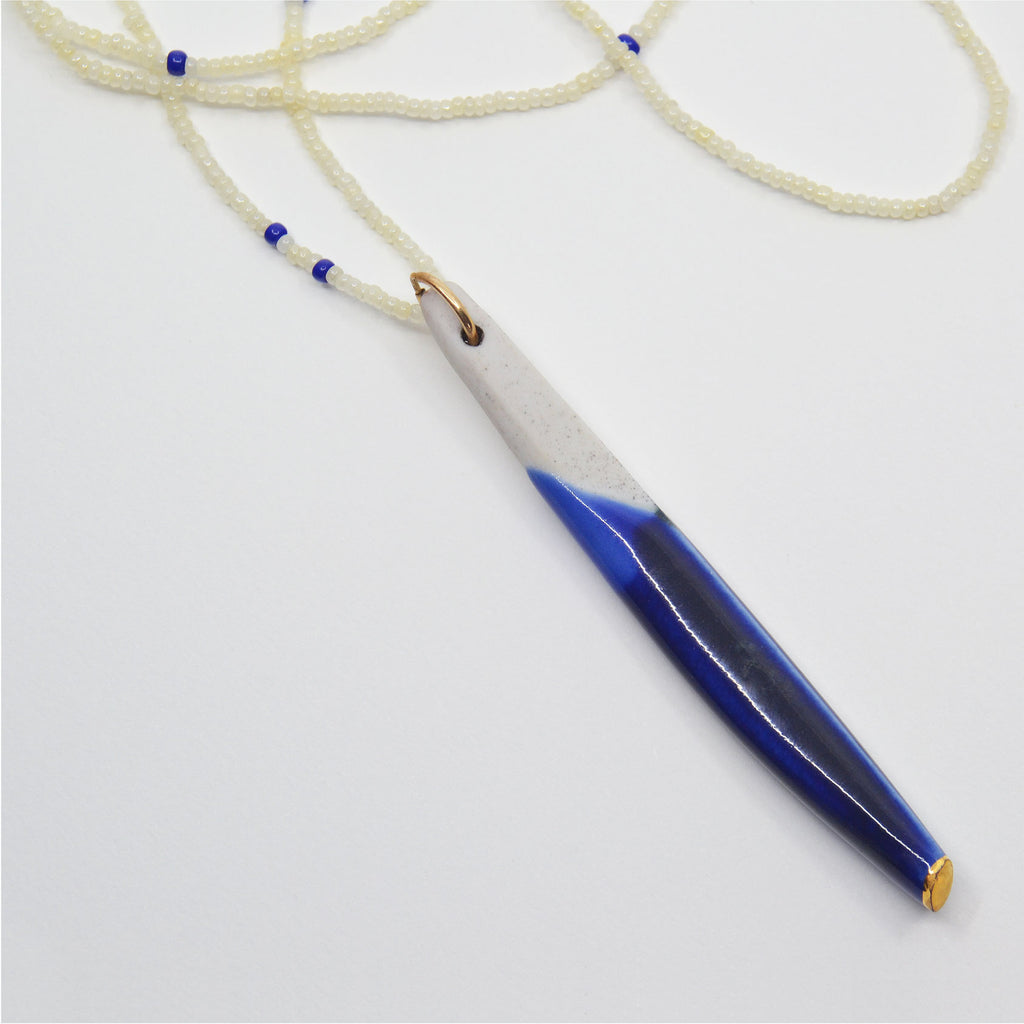 This gorgeous pendant necklace is made by hand. The pendant is hand-crafted of porcelain and shaped like an elegant long quill. The pendant is glazed with hues of blue and the tip is glazed with 22 karat gold, whereas the top is left unglazed. It is attached to a 14 karat gold-filled ring and a necklace made of tiny white vintage glass beads with a few blue beads sprinkled in-between.
