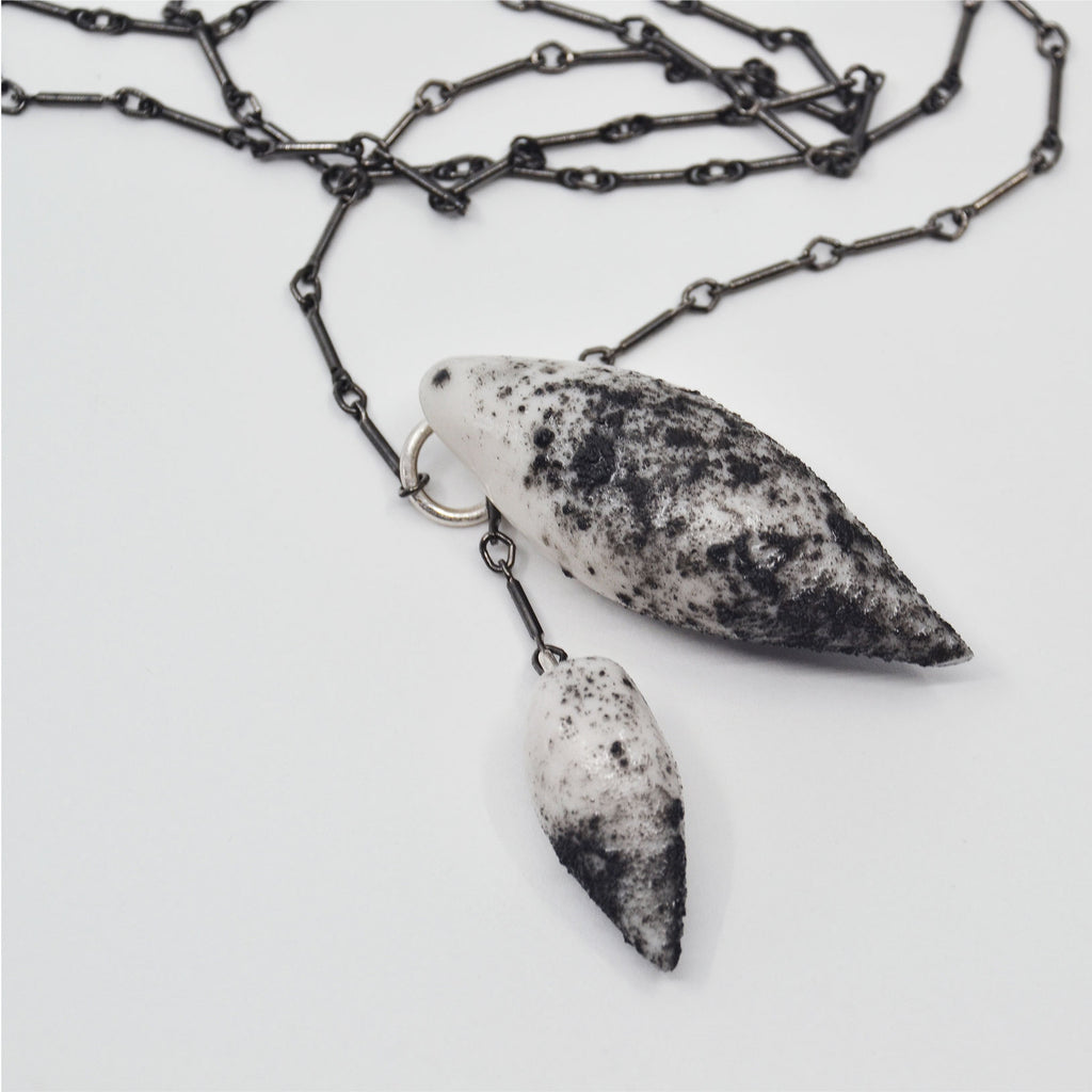 This unique pendant necklace is made by hand. It consists of two pendants crafted of white porcelain, with a black porcelain dusted surface. The pendants are attached to a sterling silver ring and gunmetal chain. 