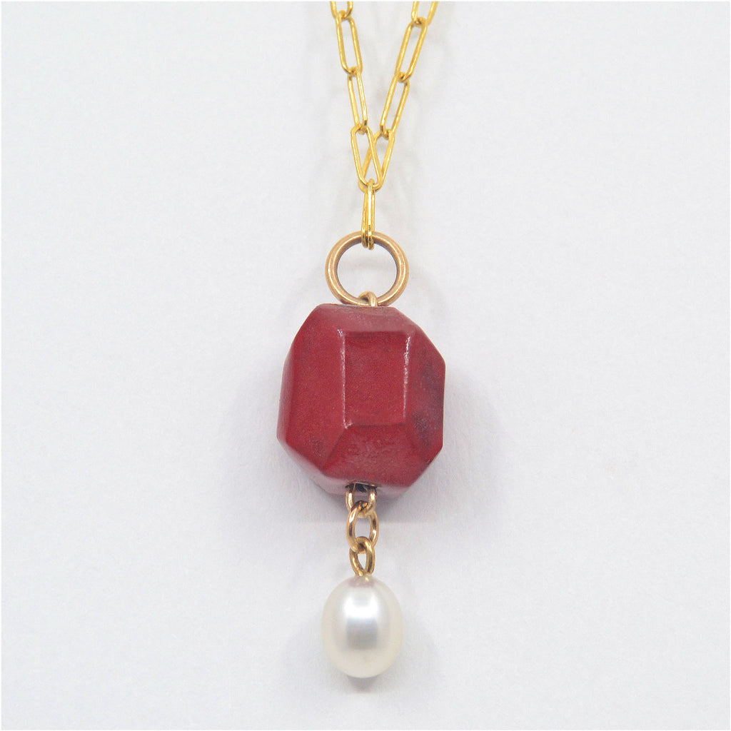 This charming unique pendant necklace is handmade. It consists of a hand-faceted porcelain bead, glazed in deep red, and is adorned with a dangling, oval shaped fresh water pearl in shimmering white. The pendant is attached to a 14 karat gold-filled ring and chain with clasp.