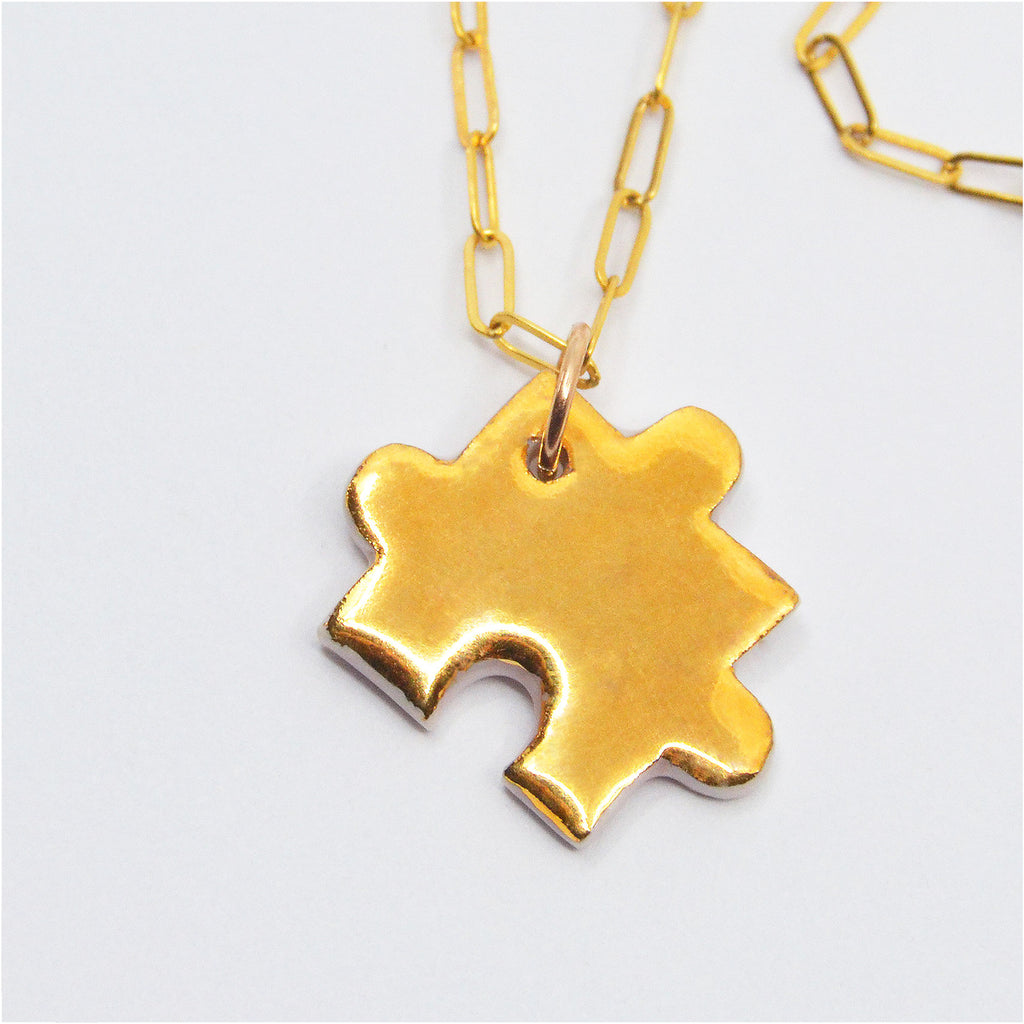 This unique pendant is handcrafted of porcelain in the shape of a puzzle piece. One side is glazed with 22 karat gold, while the other side is glossy white porcelain. The puzzle piece is attached to a 14 karat gold-filled ring and chain with clasp.