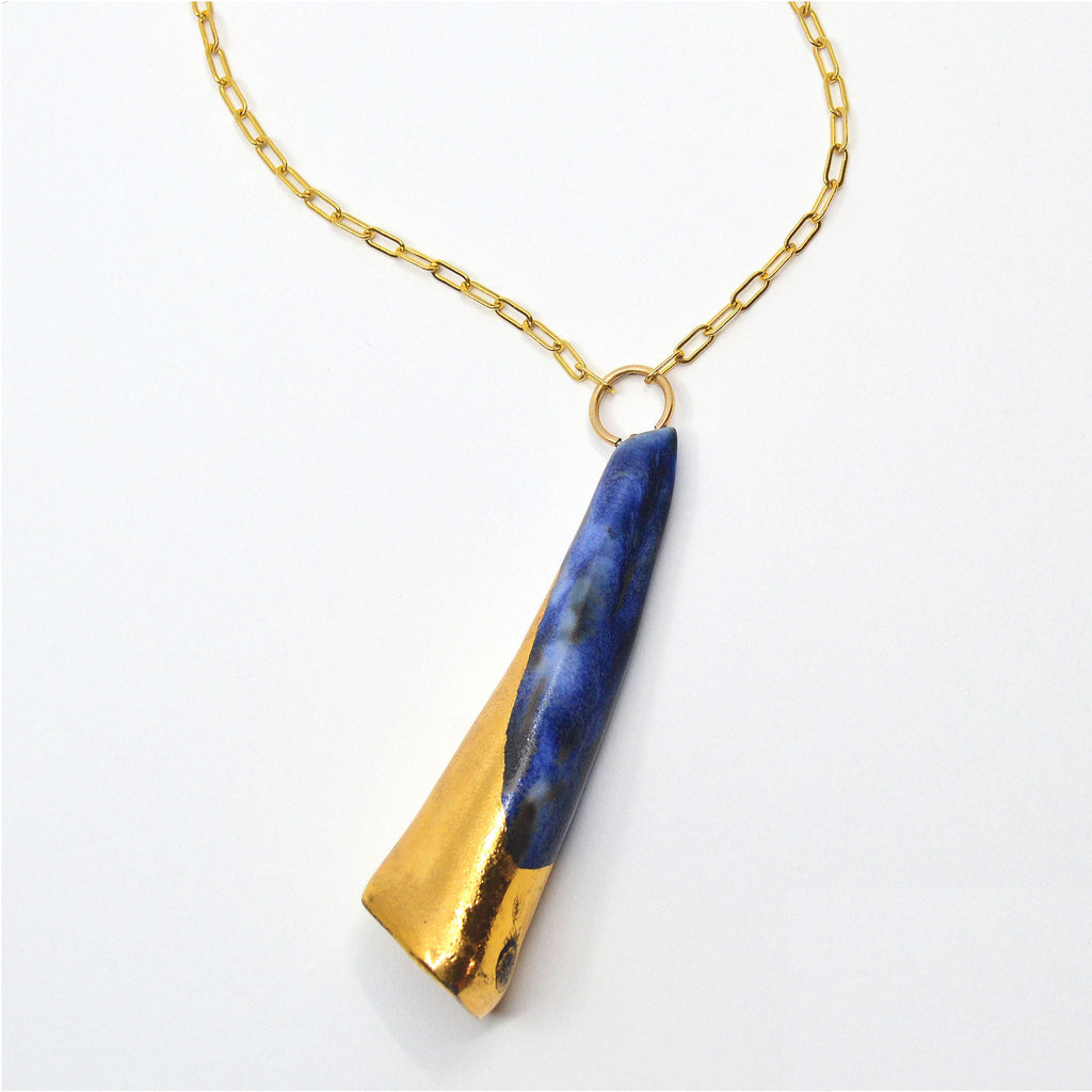 This unique pendant necklace is handmade. The pendant is handcrafted of porcelain in the shape of a cone, and glazed with irregular blue glaze with darker and lighter spots, evoking a stormy night sky. The bottom  part of the cone is glazed with 22 karat gold in an asymmetric design. The pendant is attached to a 14 karat gold-filled ring and long chain. 