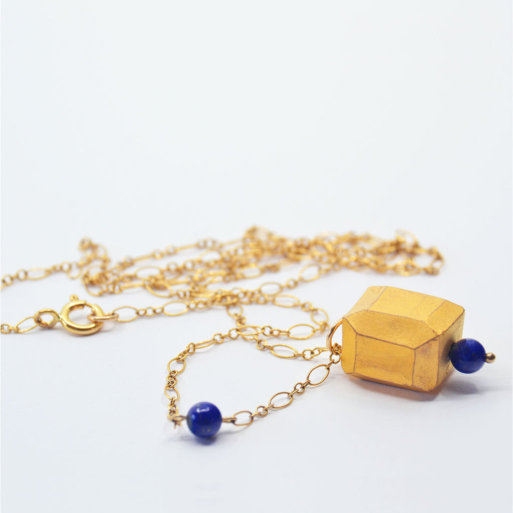 This pretty necklace is made by hand. The pendant is made of a hand-faceted porcelain bead, glazed in matte 22 karat gold, and is adorned with a lapis lazuli bead. The pendant is attached to a 14 karat gold-filled chain with clasp. There is another lapis lazuli bead along one side of the chain.