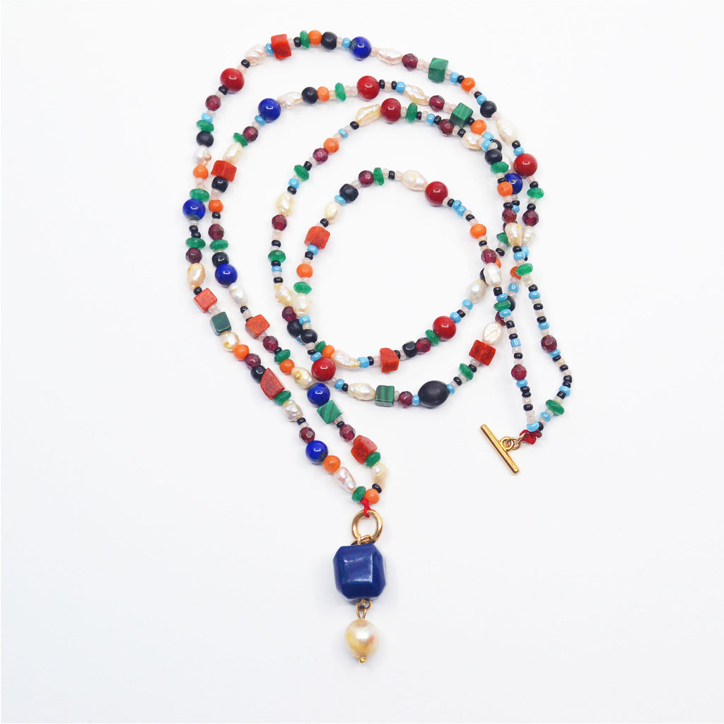 This stunning pendant necklace is made by hand. The pendant consists of a handcrafted porcelain bead, glazed in blue and a gorgeous vintage pearl. The pendant is attached to a 14 karat gold-filled ring and necklace. The necklace is made of a colorful array of vintage pearls, corals and glass beads, and lapis lazuli, emeralds, malachite and amethysts. The necklace can be worn long or as a choker with a 14 karat gold-filled toggle clasp, which closes in the front.