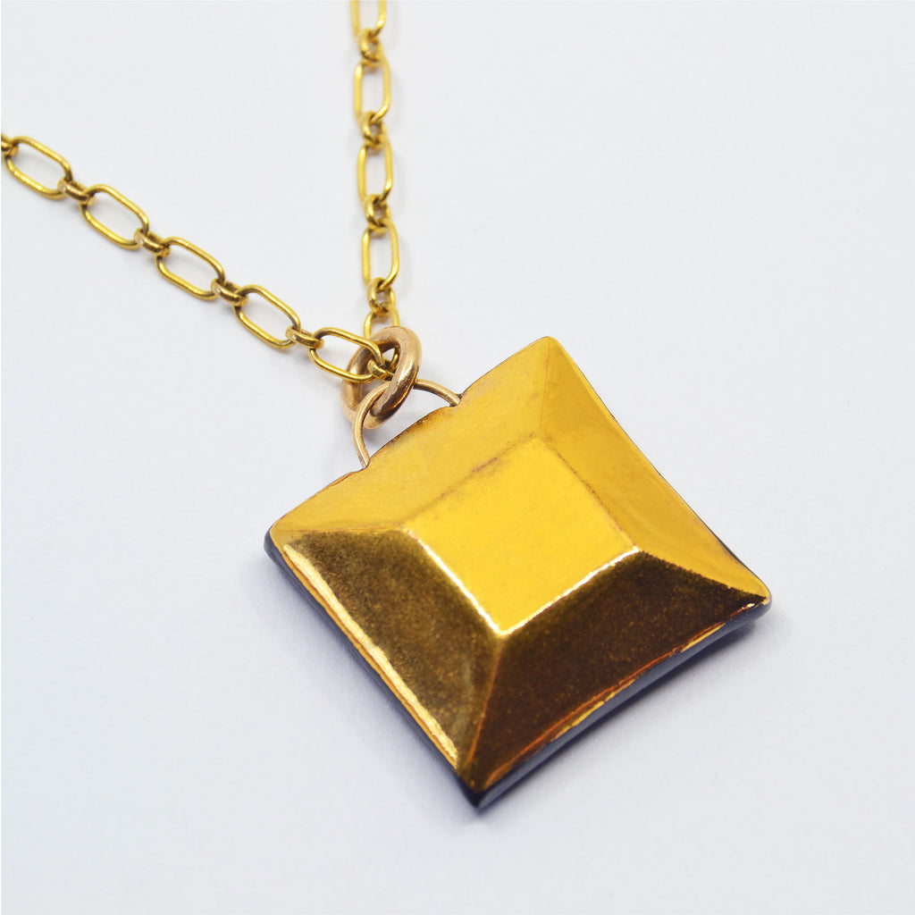 This unique pendant is handmade of black porcelain, and faceted by hand. The front is glazed with 22 karat gold, and the back of the pendant is left unglazed, creating a striking contrast between the surfaces. The pendant is attached to a 14 karat gold-filled ring and chain.