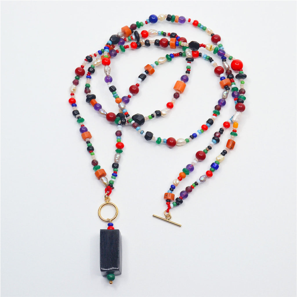 This stunning pendant necklace is made by hand. The pendant consists of a handcrafted porcelain bead, glazed in black, as well as vintage glass beads in deep red and blue, and a malachite bead. The pendant is attached to a 14 karat gold-filled ring and necklace. The necklace is made of a colorful array of vintage pearls, corals and glass beads, and lapis lazuli, emeralds and amethysts. The necklace can be worn long or as a choker with a 14 karat gold-filled toggle clasp, which closes in the front.