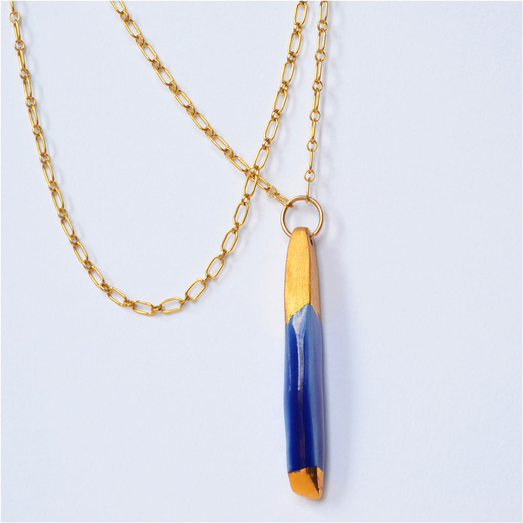 This unique pendant necklace is made by hand. The pendant is hand-crafted of porcelain and glazed with glossy blue and 22 karat gold, in both shiny and matte. The pendant is attached to a 14 karat gold-filled ring and long chain.