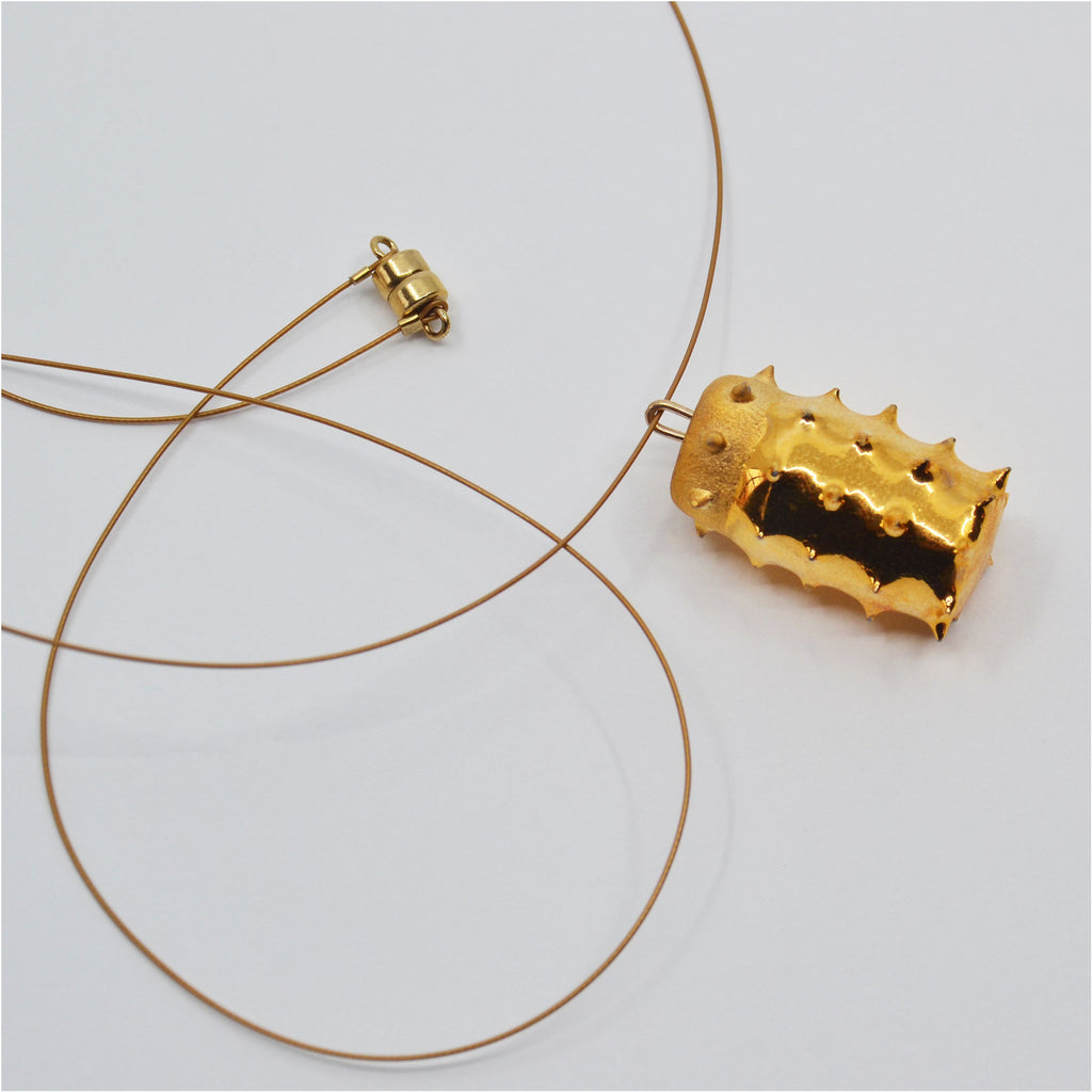 This unique necklace is handmade. The pendant is handcrafted in the shape of a cylinder with pointy spikes. It is glazed with glossy 22 karat gold and matte at the top. The pendant is attached to a golden jewelry cord with a 14 karat gold-filled magnetic clasp.