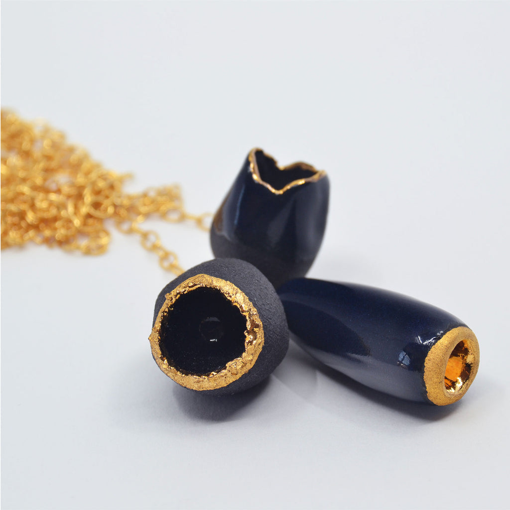 This unique pendant necklace is made by hand. The flower bud shaped beads are handcrafted of porcelain, and glazed with darkest midnight blue, black and 22 karat gold glaze, parts of the porcelain remain unglazed, creating a striking contrast between the smooth glazed texture and the raw unglazed surfaces. The beads are attached to a 14 karat gold-filled ring and long chain.
